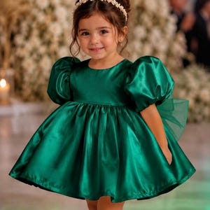 May include: A dark emerald green satin dress featuring short puff sleeves, a round neckline, and a full, pleated skirt. The dress is paired with a white pearl-embellished headband and matching dark emerald green patent leather Mary Jane shoes.