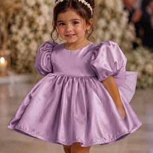 May include: A lavender satin dress featuring short puff sleeves, a round neckline, and a full, pleated skirt. The dress is paired with a white pearl-embellished headband and cream-colored flat shoes with a single strap across the top of the foot.