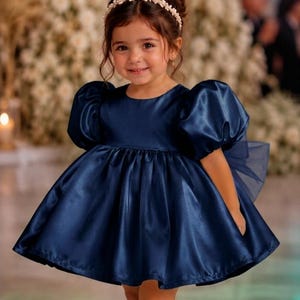 May include: A navy blue satin dress with short puff sleeves, a round neckline, and a full, pleated skirt. The dress features a sheer tulle bow at the back. The outfit is paired with a pearl-embellished headband and navy blue patent leather shoes.