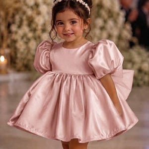 May include: A light pink satin dress featuring short puff sleeves, a round neckline, and a full, pleated skirt. The dress is paired with a white floral headband and cream-colored flat shoes with a single strap across the top of the foot.