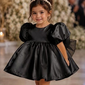May include: A black satin dress with short puff sleeves, a round neckline, and a full, pleated skirt. The dress features a large sheer bow at the back. A white pearl-embellished headband and cream-colored Mary Jane shoes complete the ensemble.