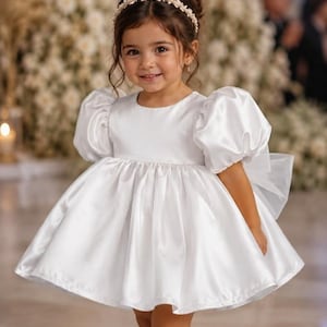 May include: A white satin dress with short puff sleeves, a round neckline, and a full, pleated skirt. The dress features a large bow on the back and is paired with white patent leather Mary Jane shoes and a pearl-embellished headband.