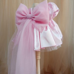 May include: A light pink dress featuring a satin bodice with short puff sleeves and a voluminous tulle skirt. A large, oversized sheer organza bow is attached to the back, with long, flowing fabric panels extending downward from the bow's center.
