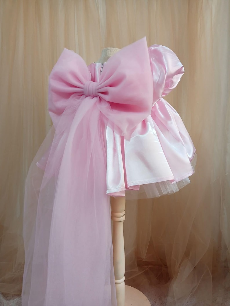 May include: A light pink dress featuring a satin bodice with short puff sleeves and a voluminous tulle skirt. A large, oversized sheer organza bow is attached to the back, with long, flowing fabric panels extending downward from the bow's center.