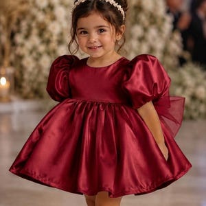 May include: A deep burgundy satin dress featuring short puff sleeves, a round neckline, and a full, flared skirt. The dress includes a large sheer bow detail at the back. The garment is paired with cream-colored Mary Jane shoes and a pearl headband.