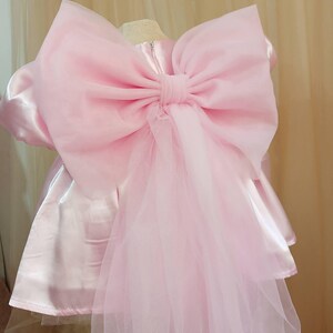 May include: A light pink satin dress featuring a large, voluminous organza bow attached to the back. The dress has short puffed sleeves and a smooth, shiny finish, with a long, sheer tulle train cascading down from the center of the oversized bow.