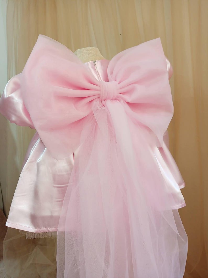 May include: A light pink satin dress featuring a large, voluminous organza bow attached to the back. The dress has short puffed sleeves and a smooth, shiny finish, with a long, sheer tulle train cascading down from the center of the oversized bow.