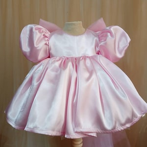 May include: A light pink satin dress featuring voluminous puff sleeves, a round neckline, and a full, gathered skirt. The garment includes a large, structured bow detail at the back, creating a silhouette with a smooth, shiny finish throughout.