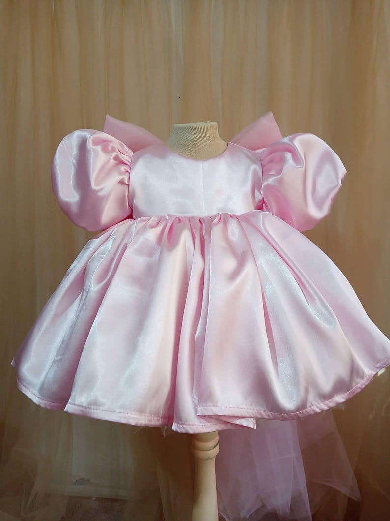 May include: A light pink satin dress featuring voluminous puff sleeves, a round neckline, and a full, gathered skirt. The garment includes a large, structured bow detail at the back, creating a silhouette with a smooth, shiny finish throughout.