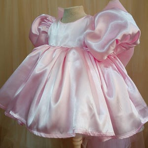 May include: A light pink satin dress featuring short, voluminous puff sleeves and a wide, gathered skirt. The garment has a round neckline and a large, prominent bow attached to the back, creating a structured silhouette with a smooth, shiny finish.