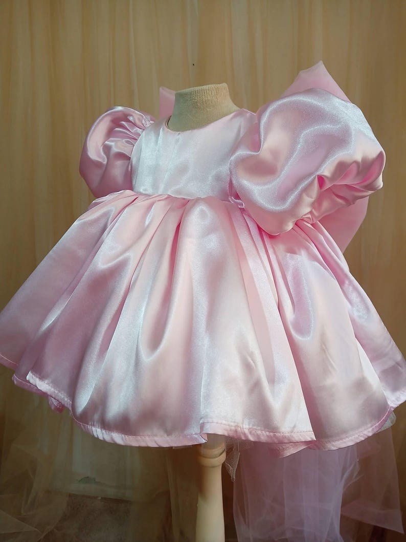 May include: A light pink satin dress featuring short, voluminous puff sleeves and a wide, gathered skirt. The garment has a round neckline and a large, prominent bow attached to the back, creating a structured silhouette with a smooth, shiny finish.