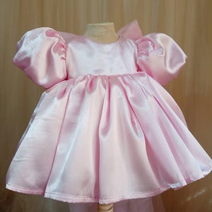 May include: A light pink satin dress featuring short, voluminous puff sleeves, a round neckline, and a gathered, full skirt that flares outward. The garment has a smooth, shiny finish and a structured bodice, displayed on a mannequin form.