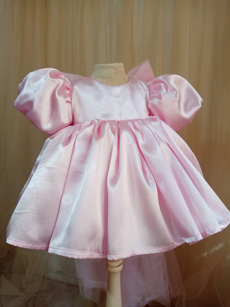 May include: A light pink satin dress featuring short, voluminous puff sleeves, a round neckline, and a gathered, full skirt that flares outward. The garment has a smooth, shiny finish and a structured bodice, displayed on a mannequin form.