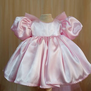 May include: A light pink satin dress featuring voluminous puff sleeves, a round neckline, and a full, pleated skirt. The garment has a structured bodice and a large bow detail visible at the back, showcasing a smooth, reflective fabric finish.