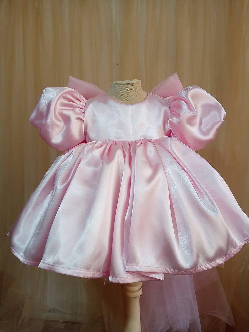 May include: A light pink satin dress featuring voluminous puff sleeves, a round neckline, and a full, pleated skirt. The garment has a structured bodice and a large bow detail visible at the back, showcasing a smooth, reflective fabric finish.