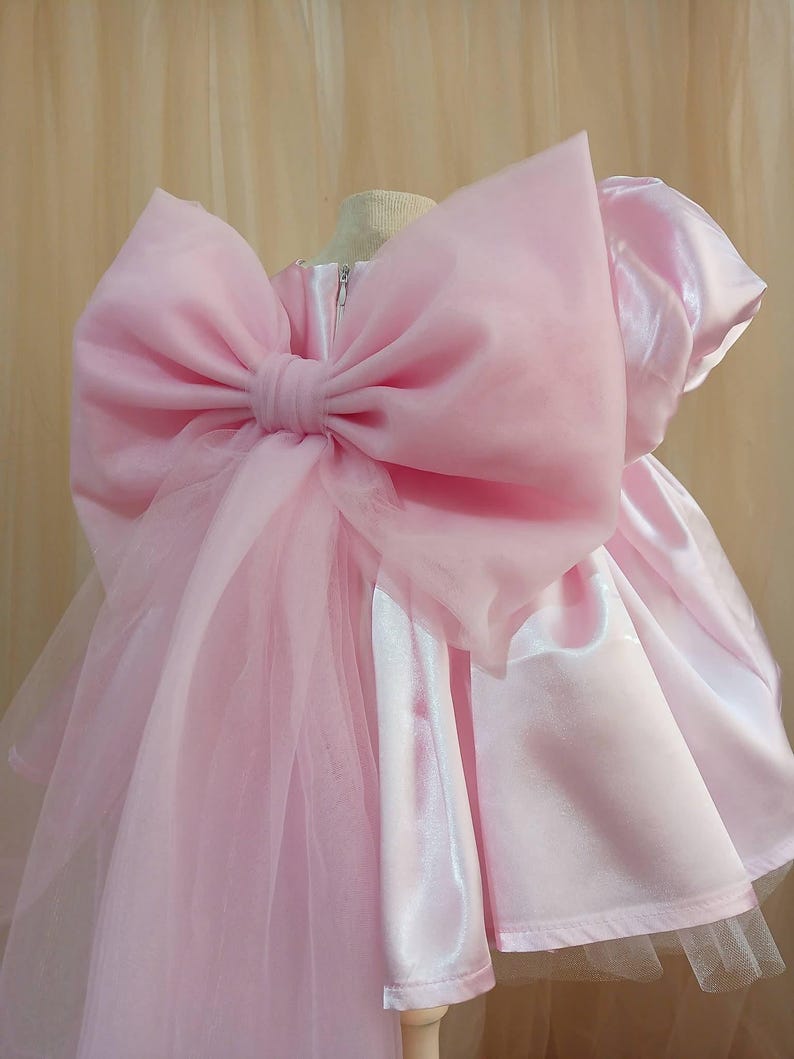 May include: A light pink dress featuring a large, sheer organza bow attached to the back. The dress is constructed from a smooth, satin-like fabric with short sleeves and a zipper closure visible at the center back, accented by layers of tulle.