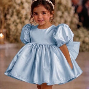 May include: A light blue satin dress featuring short puff sleeves, a round neckline, and a full, pleated skirt. The dress includes a large bow detail on the back and is paired with cream-colored flat shoes and a floral headband with small white flowers.