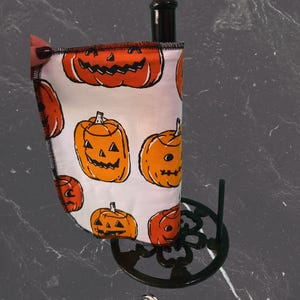 May include: A Halloween wine bottle cooler with a white fabric covered in orange pumpkin designs. The cooler is held by a black metal stand. The cooler is wrapped around a black bottle. The image includes the text "Izaban Creations".