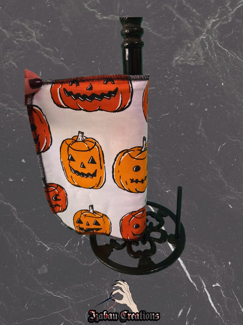 May include: A Halloween wine bottle cooler with a white fabric covered in orange pumpkin designs. The cooler is held by a black metal stand. The cooler is wrapped around a black bottle. The image includes the text "Izaban Creations".