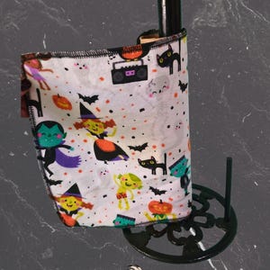 May include: A black bottle with a Halloween-themed fabric wrap featuring cartoon witches, bats, and pumpkins. The fabric is wrapped around the bottle and held by a black metal stand. The fabric has a white background with colourful illustrations.