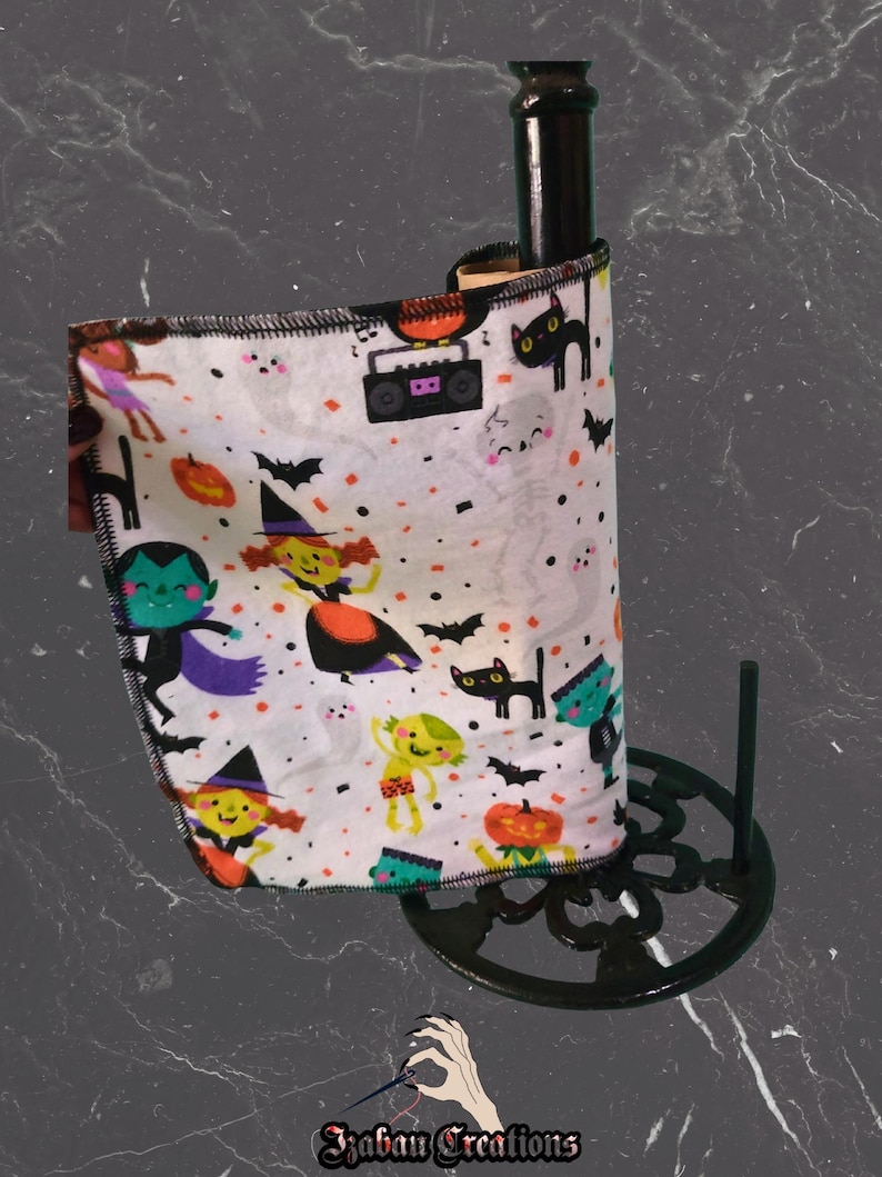 May include: A black bottle with a Halloween-themed fabric wrap featuring cartoon witches, bats, and pumpkins. The fabric is wrapped around the bottle and held by a black metal stand. The fabric has a white background with colourful illustrations.