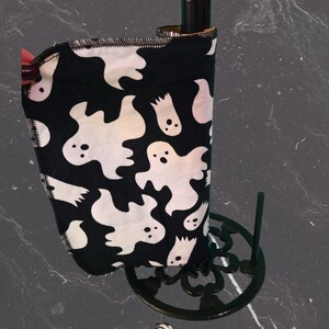 May include: A black and white ghost-themed paper towel holder. The fabric features white ghost illustrations on a black background. The holder is on a black metal stand. The brand name "Izaban Creations" is at the bottom.