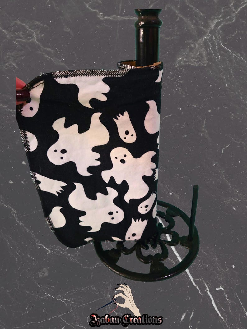 May include: A black and white ghost-themed paper towel holder. The fabric features white ghost illustrations on a black background. The holder is on a black metal stand. The brand name "Izaban Creations" is at the bottom.