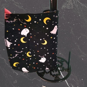 May include: Black wine bottle sleeve with a Halloween design. The sleeve displays white ghosts, yellow crescent moons, and stars on a black backdrop. The bottle is held in a decorative black metal stand.