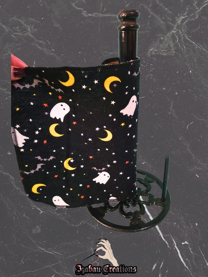 May include: Black wine bottle sleeve with a Halloween design. The sleeve displays white ghosts, yellow crescent moons, and stars on a black backdrop. The bottle is held in a decorative black metal stand.