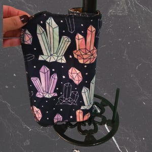 May include: A black wine bottle cooler featuring a geometric crystal design in pink, green, and orange. The cooler is held by a black metal stand. The text "Izaban Creations" is at the bottom of the image.