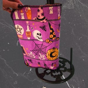 May include: A purple paper towel holder with a black base, holding a purple paper towel with Halloween illustrations. The towel features witch hats, candles, skulls, and pumpkins. The base has an openwork design.