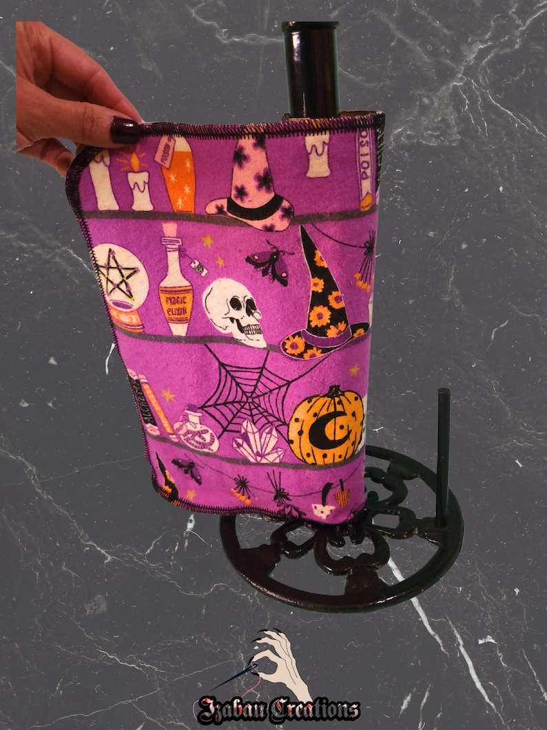 May include: A purple paper towel holder with a black base, holding a purple paper towel with Halloween illustrations. The towel features witch hats, candles, skulls, and pumpkins. The base has an openwork design.
