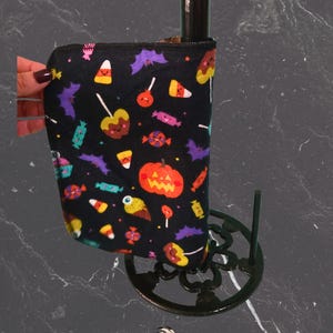 May include: Black Halloween fabric sleeve featuring candy corn, bats, and pumpkin motifs. The sleeve is wrapped around a dark brown bottle. The sleeve is held by a hand with dark nail polish. The base is a decorative metal stand.