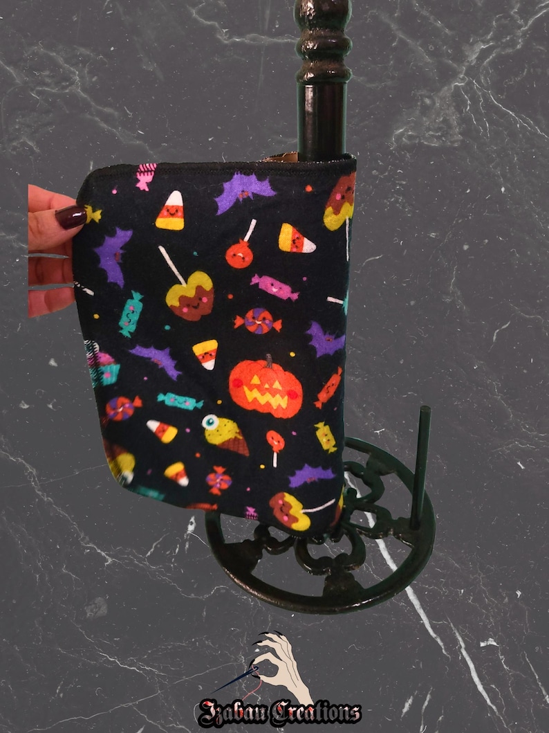 May include: Black Halloween fabric sleeve featuring candy corn, bats, and pumpkin motifs. The sleeve is wrapped around a dark brown bottle. The sleeve is held by a hand with dark nail polish. The base is a decorative metal stand.