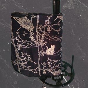 May include: A black and white decorative bottle holder featuring a spooky tree and cat design. The holder wraps around a dark brown bottle and rests on a black metal base. The design includes a bat, a cat, and the text "Izaban Creations".