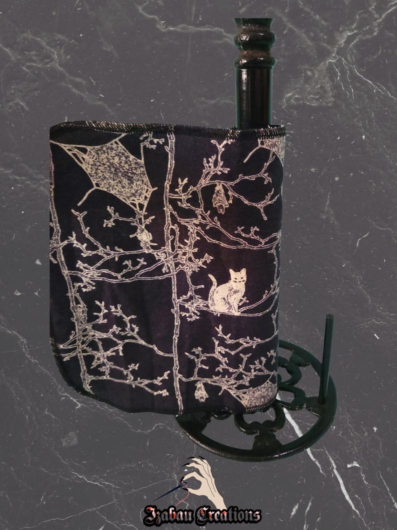 May include: A black and white decorative bottle holder featuring a spooky tree and cat design. The holder wraps around a dark brown bottle and rests on a black metal base. The design includes a bat, a cat, and the text "Izaban Creations".