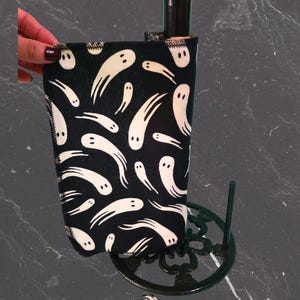 May include: A black wine bottle sleeve featuring a white ghost design. The sleeve is wrapped around a dark bottle, set in a dark green metal stand. The image includes the text "Izaban Creations".