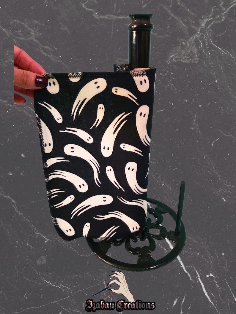 May include: A black wine bottle sleeve featuring a white ghost design. The sleeve is wrapped around a dark bottle, set in a dark green metal stand. The image includes the text "Izaban Creations".