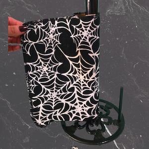 May include: A black and white spiderweb-patterned wine bottle cooler. The cooler is wrapped around a dark bottle, held by a decorative black metal stand. The cooler is made of fabric and is designed to keep wine chilled.