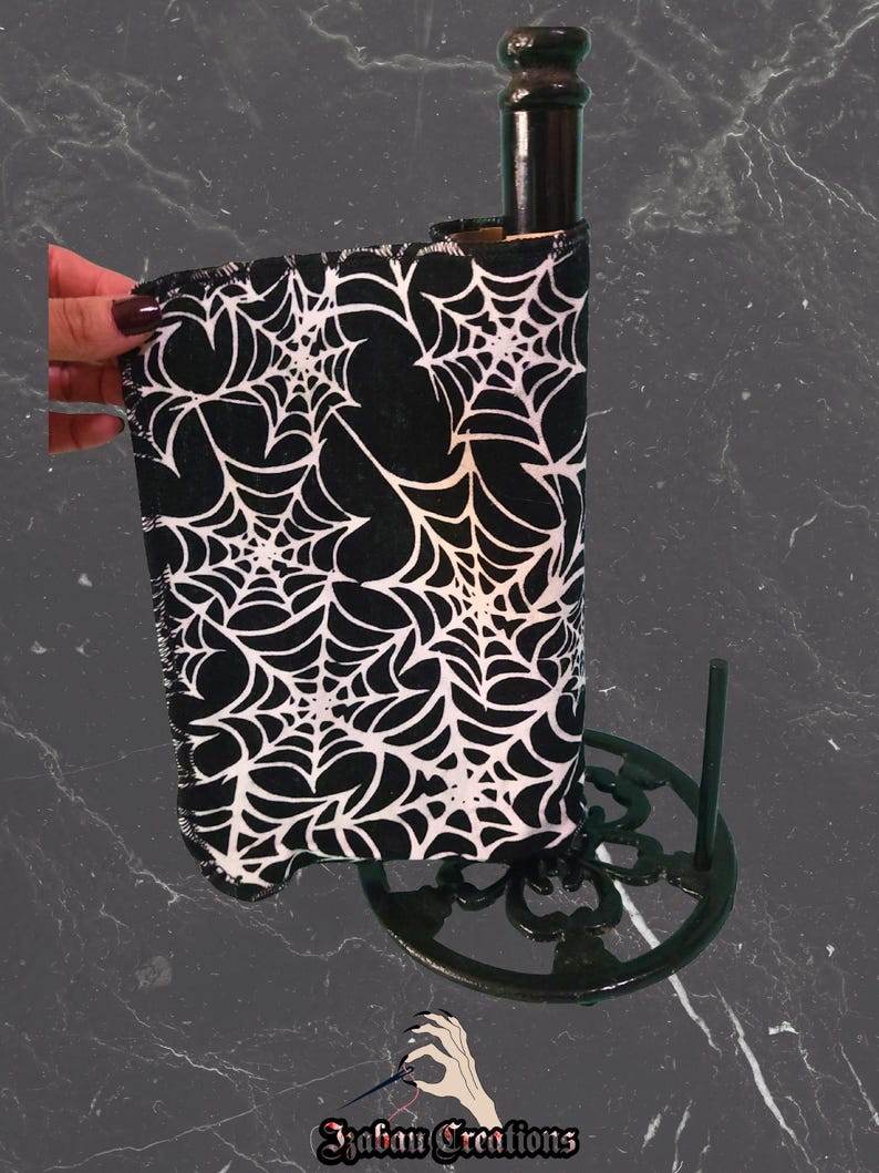May include: A black and white spiderweb-patterned wine bottle cooler. The cooler is wrapped around a dark bottle, held by a decorative black metal stand. The cooler is made of fabric and is designed to keep wine chilled.