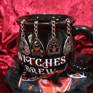May include: Black ceramic mug with the words "Witches Brew" in white lettering. The mug has a black handle and is adorned with silver-colored planchette charms hanging from the rim. The charms have various symbols and the words "Yes" and "No".