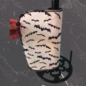 May include: A white fabric wine bottle cooler featuring a black bat pattern. The cooler is wrapped around a black bottle, and the design is set on a black metal stand. The edges of the cooler are stitched with black thread.