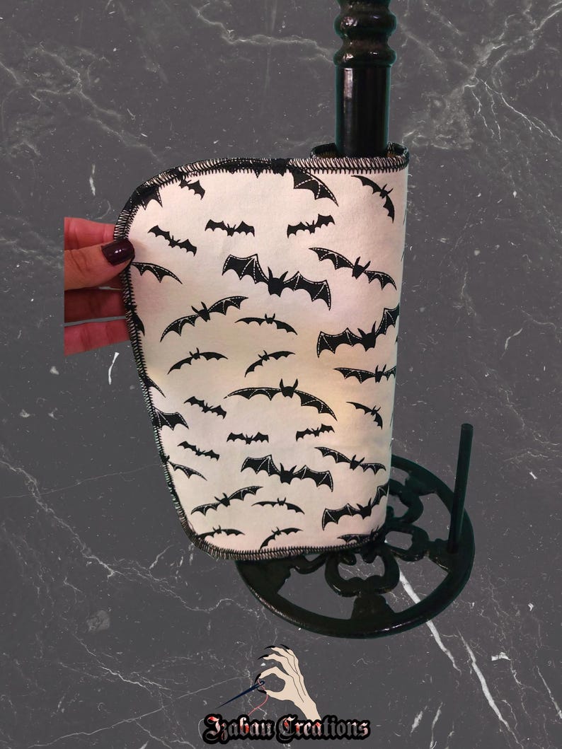 May include: A white fabric wine bottle cooler featuring a black bat pattern. The cooler is wrapped around a black bottle, and the design is set on a black metal stand. The edges of the cooler are stitched with black thread.