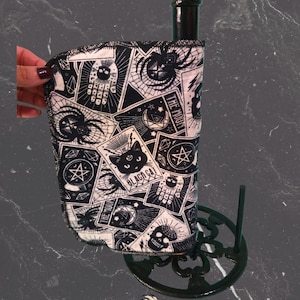 May include: Black and white paper towel holder with a decorative print featuring tarot card imagery, skulls, spiders, and a black cat. The holder is made of metal and has a dark finish. The paper towel roll is partially visible.