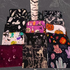 May include: A selection of Halloween-themed fabric items, including bottle covers and small cloths. The bottle cover is white with black bat silhouettes. The cloths feature various spooky designs like ghosts, pumpkins, and crystals. The color palette includes black, white, purple, and orange.