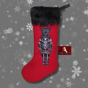 May include: A red Christmas stocking featuring a black faux fur cuff and a graphic print of a skeleton nutcracker wearing a top hat. The nutcracker has red eyes and a white beard. A small rectangular logo reads Izabau Creations in white text.