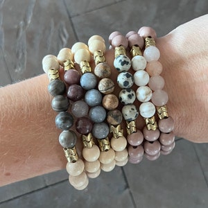 May include: A stack of eight bracelets made with various natural stones and gold accents. The bracelets feature different colors and patterns, including brown, beige, gray, white, and pink.