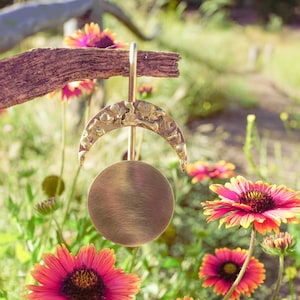 Puede incluir: Un carillón de viento de color latón con una luna creciente y un disco circular. El carillón cuelga de una rama de madera sobre un fondo de flores coloridas y vegetación. La luna tiene una superficie texturizada.
