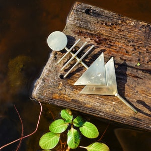 May include: A collection of brass geometric shapes, including a circle, triangles, and linear elements, arranged on a weathered wooden plank. The objects are set against a backdrop of water and greenery, creating a natural, artistic composition.