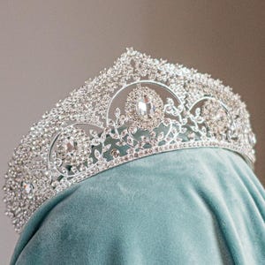May include: A silver tiara with numerous clear crystals and gemstones. The crown is set on a teal fabric, with a detailed filigree design and large central stones. The tiara has a classic, elegant design.